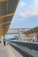 Ratchaburi Railway Station in Thailand, featuring dual train tracks, golden-yellow platform, and striking overhead crossing. Clear blue sky with soft clouds sets a perfect travel mood. photo
