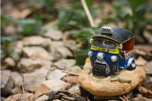 Small colorful tuk-tuk model made from painted clay, arranged in a rural vintage setting. Positioned right with empty space for text on the left. Natural light, nostalgic countryside vibe. photo