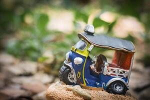Close-up of a miniature Thai tuk-tuk sculpture with a visible flat tire, centered in a vintage countryside setting. Soft-focus small trees behind create an old-world nostalgic atmosphere. photo