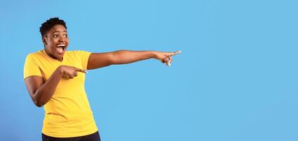 Look There. Excited Overweight African Lady Pointing Fingers Aside At Free Space For Text Standing Over Blue Background. Advertisement Banner. Panorama, Studio Shot photo