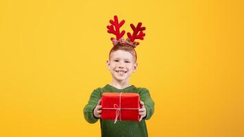 Smiling boy with festive deer accessory giving Christmas gift over yellow studio background, empty space. Happy kid celebrating New Year, cheerfully outstretching present to camera photo