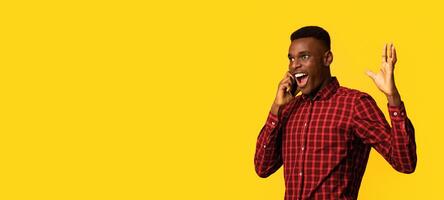 Mobile Communication. Excited African American Guy Emotionally Talking On Cellphone, Having Pleasant Phone Conversation, Standing Isolated Over Yellow Background, Panorama With Copy Space photo