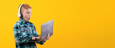Excited redhead boy with wireless headset playing on laptop, yellow studio background. Amazed kid in casual outfit using modern powerful gadget for games, playing online, copy space photo
