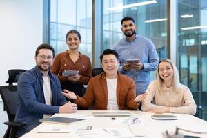 A diverse team smiles at the camera while gathered around a table in a modern office setting. Collaboration and teamwork are evident. photo
