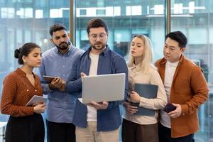 A diverse group of business professionals collaborates in an office setting, examining a laptop screen intently during a meeting. photo