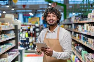 A smiling grocery store worker holds a tablet while standing in an aisle, surrounded by products on shelves. photo