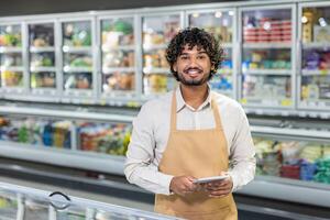 A smiling man wearing an apron holds a tablet while standing in a grocery store. photo
