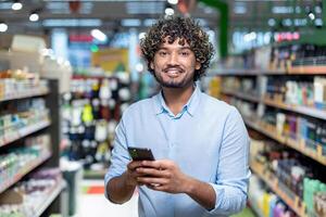 A smiling man in a supermarket, holding a phone, looking at the camera. Shelves with products are in the background. photo