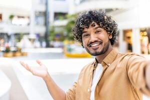 A smiling man with curly hair takes a selfie outside, extending his arm towards the viewer, invitingly. photo