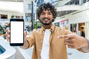 A smiling man with curly hair holds up a phone with a blank screen, pointing at it in a modern mall. photo