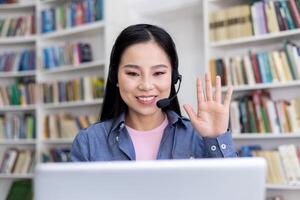 A smiling Asian woman wearing a headset waves during a call in front of a bookshelf. photo