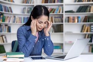 A young Asian woman experiencing a headache while studying in a library, surrounded by books and using a laptop. photo