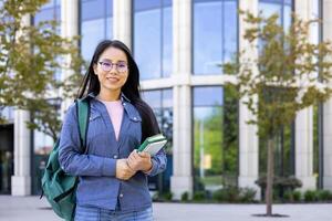 A smiling Asian student with glasses stands in front of a university building, holding books and a backpack. photo