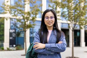A smiling Asian woman wearing glasses stands with her arms crossed in front of a modern building. photo