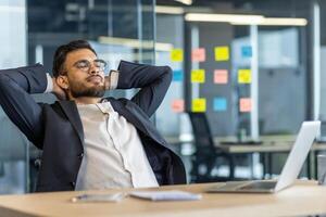 Businessman relaxing and resting inside the office. Man in business suit with hands behind head with closed eyes thinking and dreaming. Boss at workplace, working with laptop. photo