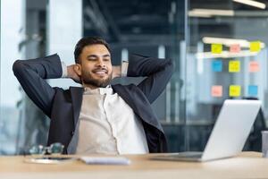 A smiling businessman in a suit relaxes in his office, leaning back with hands behind his head, enjoying his work. photo