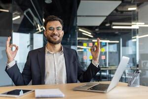 A smiling businessman sits at his desk in the office, gesturing with his hands in a zen-like expression. He appears calm and focused, embracing a moment of mindfulness photo