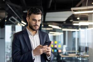 A focused businessman in a suit checks his smartphone, the modern office blurred in the background. He appears deep in thought while reading messages or emails. photo