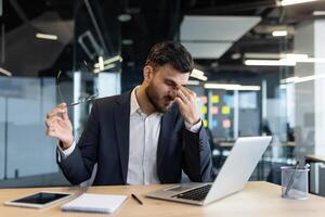 A businessman in a suit, experiencing a headache, takes off his glasses at his desk. He is using a laptop, likely working hard, and is feeling stressed. photo