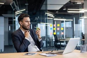 Man suffering from sore throat, company employee using spray inhaler medicine for relief. Businessman in business suit working inside office with laptop. photo