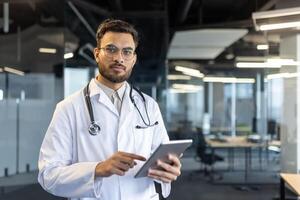 Portrait of serious thoughtful doctor, man in white medical coat and with stethoscope looking at camera. Medic uses tablet computer for online communication with patients. photo