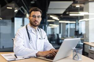 A confident doctor sits at his desk, working on a laptop in a modern office setting. He is wearing a stethoscope and looking at the camera. photo