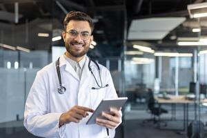 A smiling doctor in a white coat looks at the camera, holding a tablet computer. The modern office setting gives the image a professional feel. photo