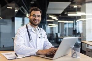 A smiling doctor, wearing a white coat and stethoscope, works on a laptop at a desk in a modern office setting. photo