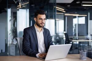 Serious thinking businessman working with laptop inside office. Man in business suit sitting at desk typing on keyboard, working late into the night. photo