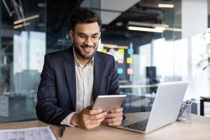 Successful businessman using tablet computer inside office at workplace. Man smiling happily, reading online financial report. photo