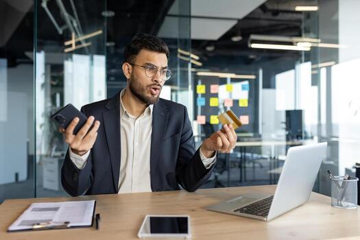 A businessman is perplexed while holding a credit card and phone. He appears to be encountering issues while using his laptop in the office. photo