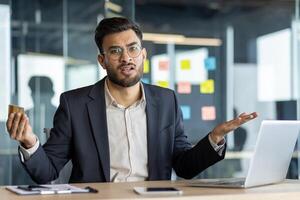 A confused businessman looks at a credit card in his hand while working on a laptop in an office environment. The man is expressing financial uncertainty. photo