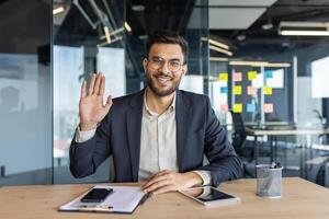 A smiling businessman waves from his desk, possibly in a conference, inside of a modern office setting. He wears a suit and glasses. photo