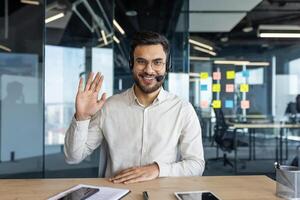 Webcam view, man with headset looking at camera, waving hello. Support service worker consultant at workplace inside office, using call. photo