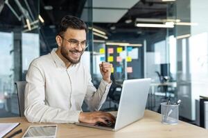 Man celebrating victory and successful achievement results at workplace. Businessman holding hand up happy smiling triumph gesture. Worker inside office working with laptop. photo