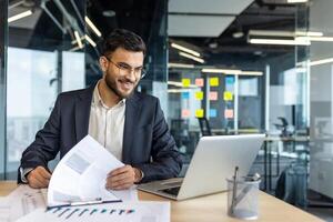 A smiling businessman reviews documents, with a laptop open on his desk in a modern office setting, ready to work. photo