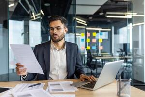 A thoughtful businessman reviews documents and uses his laptop, working in a modern office setting with a glass-walled conference room in the background photo