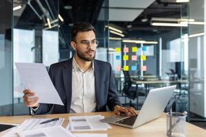 A focused businessman in glasses reviews documents while working on a laptop at his desk. The office setting features glass walls and sticky notes. photo