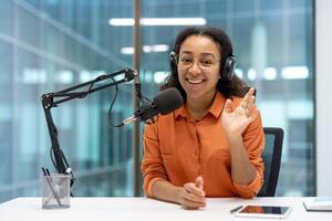 A smiling woman with glasses wearing a headset sits at a table with a microphone, recording a podcast in a bright, modern office. photo
