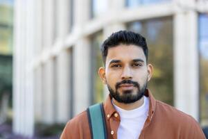 A young man with a beard and a backpack stands in front of a building on a sunny day. photo