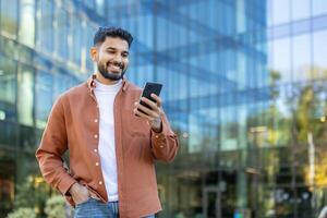 A smiling man of Indian descent looks at his phone while standing outside a modern building during the day. photo