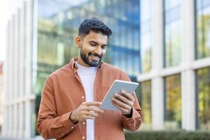 A smiling man with a beard uses a tablet outside a modern building on a sunny day, looking at the screen. photo