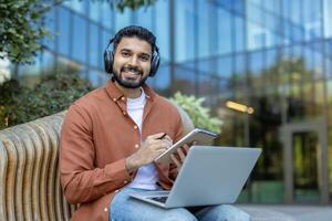 A smiling Indian man wearing headphones works outside with a laptop and notebook. He's in front of a modern building. photo