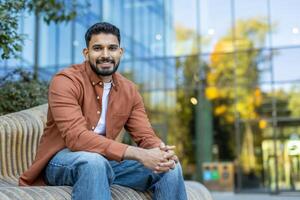 A smiling man is sitting on a bench with a modern building in the background, looking directly at the camera. photo