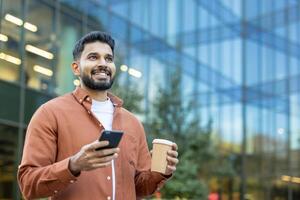A smiling Indian man holds a coffee cup and smartphone in front of a modern glass building on a sunny day. photo