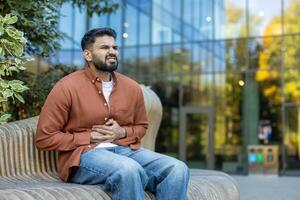A man, seated outdoors, clutches his stomach, displaying discomfort, suggesting potential illness or pain. The setting includes a bench and a modern building in the background. photo