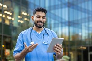 A smiling male doctor in scrubs uses a tablet outside a modern building, likely in a call. He gestures with his hands photo