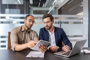 Two businessmen reviewing a tablet and laptop, discussing business strategies in a modern office setting. photo