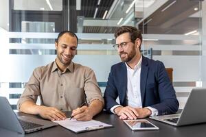 Two businessmen are sitting at a table, one is signing a document, while the other watches. They seem happy. photo