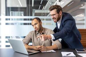 An angry manager points at a laptop while yelling at a frustrated employee in an office setting, conveying workplace stress. photo